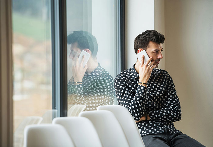 Man on phone, wearing a polka dot shirt, reflecting in a window, discussing plans to buy a house.