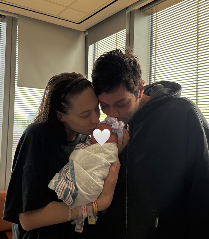 Pete Davidson and partner tenderly holding and kissing their newborn daughter in a softly lit room by window blinds.