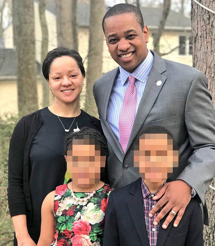 Former Virginia Lt Governor Justin Fairfax smiling outdoors with his family, blurred faces, in a casual group portrait.