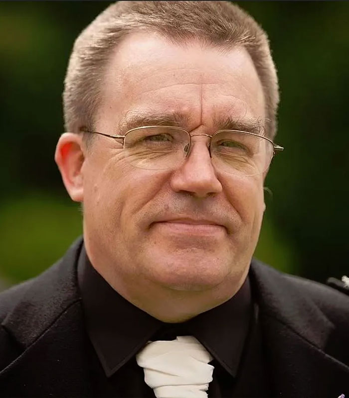 Middle-aged man wearing glasses and a black suit with white cravat, reflecting a devastated bride-to-be exposing fianc&eacute;'s crimes.