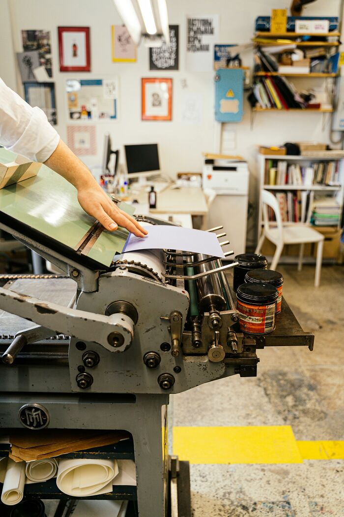 A person's hand guides paper into a letterpress machine in a bustling workplace, reminiscent of past workplace incidents.