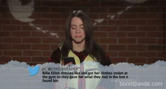 Billie Eilish reacting to a mean tweet that hits hard, showcasing a painful burn, in front of a brick wall.