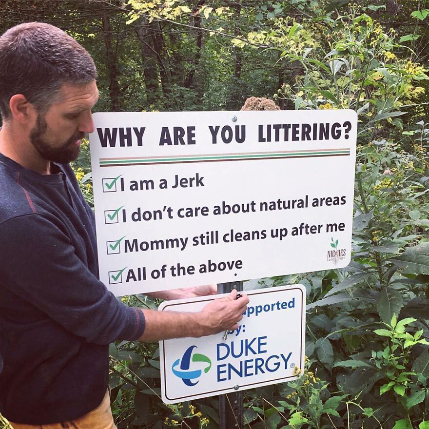 Man installing a humorous sign about littering in a forest area, highlighting camping and hiking memes about nature care.