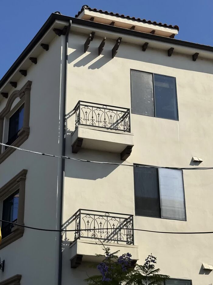 A building with balconies, ornate railings, and a terracotta roof. Shadows create an interesting, weird image on the cream wall.