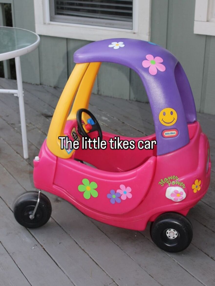 A pink, purple, and yellow Little Tikes car with flower stickers on a wooden porch, evoking growing up in the 2000s.