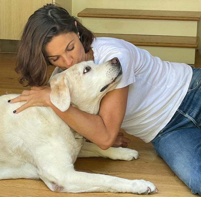 Uma mulher de camiseta branca e jeans abraçando e beijando um grande cachorro branco. O amor secreto de Mel Gibson.