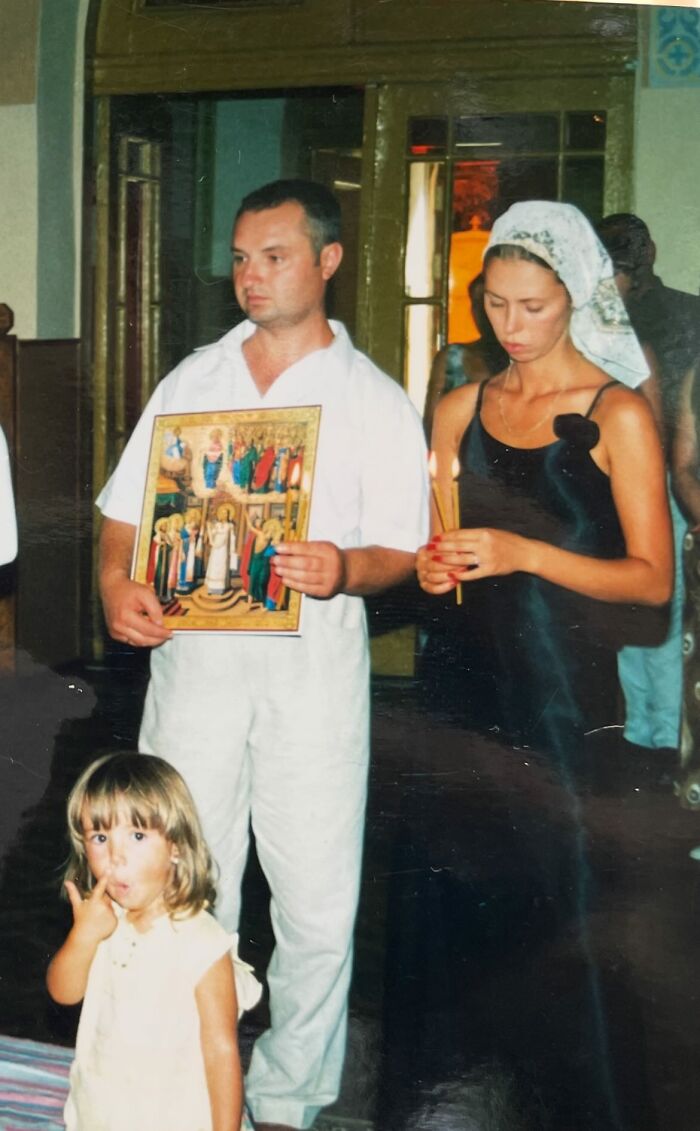 A funny and chaotic childhood photo of a girl making a silly face while adults hold religious items inside a building.