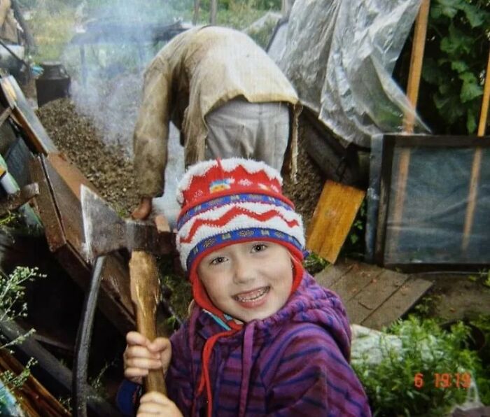 Child holding an axe and smiling outdoors with chaotic background, capturing funny and chaotic childhood moments.