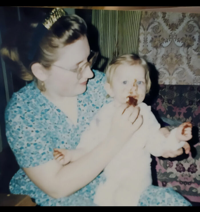 Vintage chaotic childhood photo of a toddler with food on face sitting on a woman’s lap indoors.