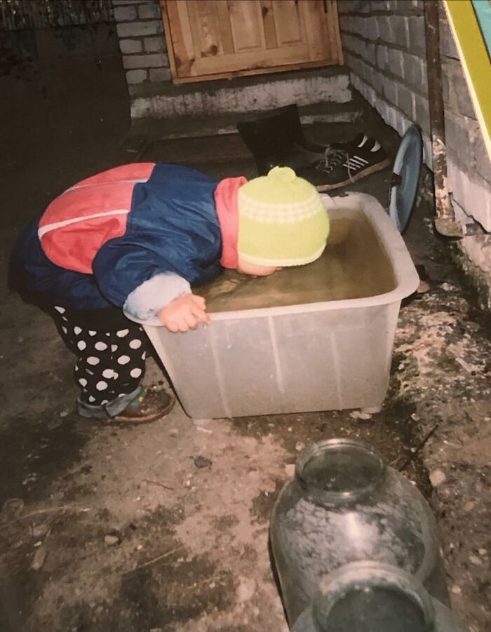 Toddler in colorful outfit drinking from large plastic container filled with water in a chaotic childhood photo moment.