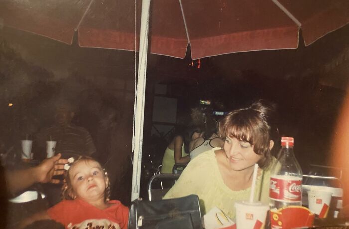 Childhood photo of a young girl making a funny face under a red umbrella at a fast food outdoor seating area.