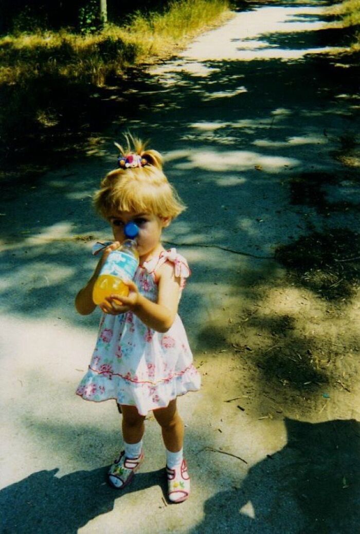 Child holding a bottle and wearing a dress outdoors in a funny and chaotic childhood photo on a sunlit path.