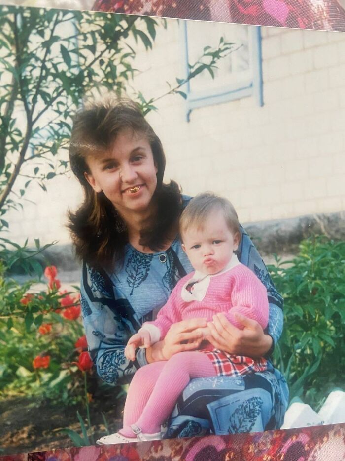 Woman in patterned dress holding toddler in pink sweater outdoors in a funny and chaotic childhood photo.