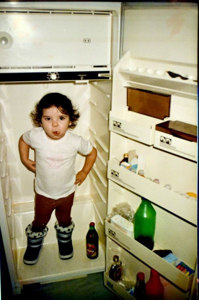 Toddler in oversized boots standing inside open refrigerator in a funny and chaotic childhood photo moment.