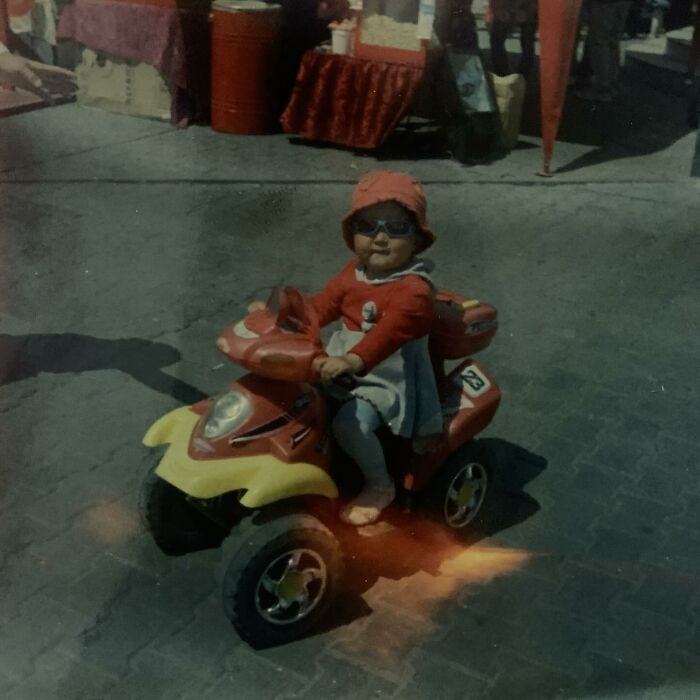 Toddler wearing sunglasses and a red hat, riding a toy ATV in a funny and chaotic childhood photo.