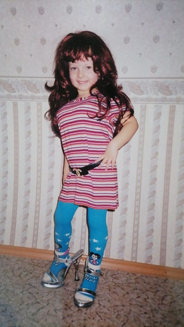 Child wearing oversized wig, striped dress, blue cartoon leggings, and high heels in a funny chaotic childhood photo.