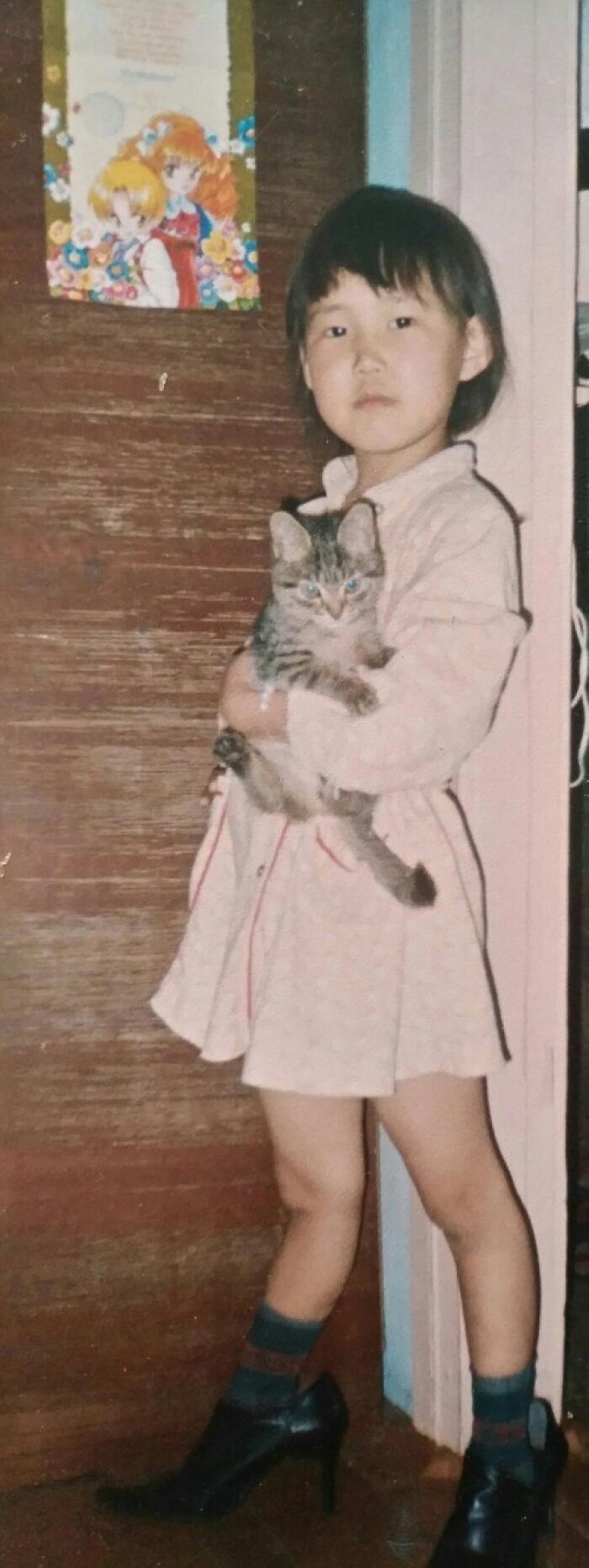 Child holding a cat wearing oversized high heels indoors, a funny and chaotic childhood photo moment.