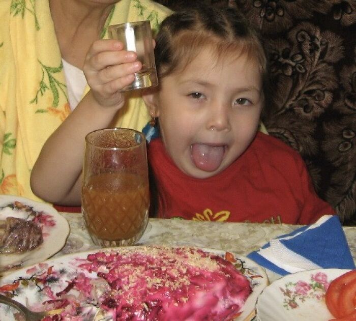 Child making a funny face at a chaotic family meal with colorful food and drinks on the table.