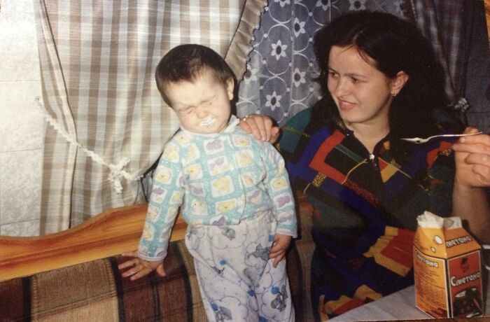 Toddler making a funny face while a woman in colorful clothes feeds him, capturing chaotic childhood photo moments.