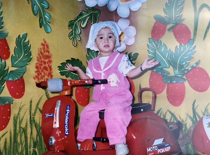 Toddler in pink overalls and bonnet sitting confused on a red vintage toy scooter in a chaotic childhood photo setting.
