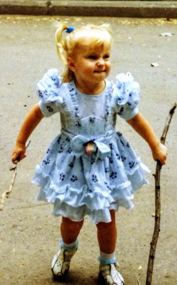 Child holding two sticks wearing a frilly dress, captured in a funny and chaotic childhood moment outdoors.