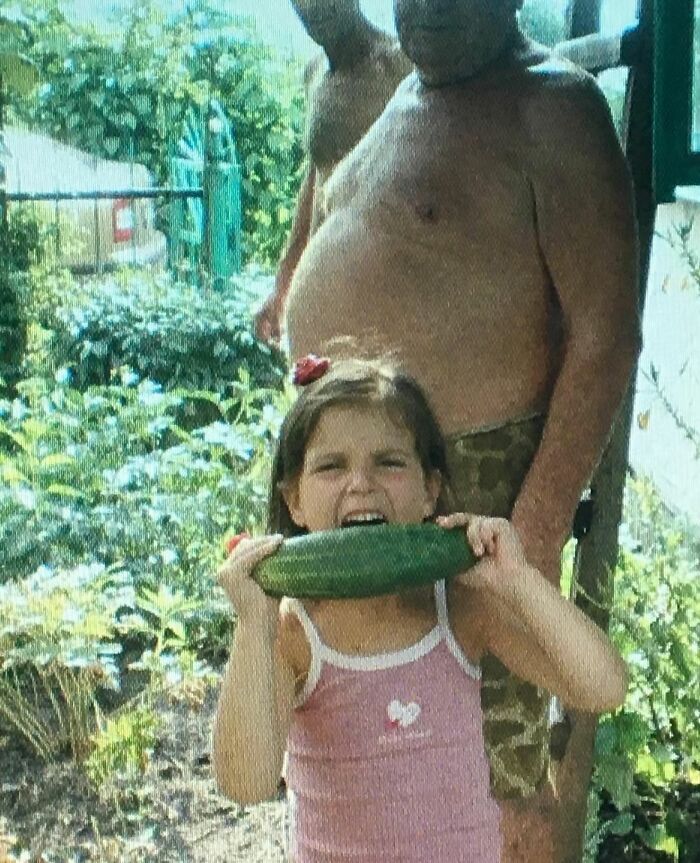 Young girl biting a large cucumber with a shirtless man in camouflage shorts in a chaotic childhood photo outdoors.