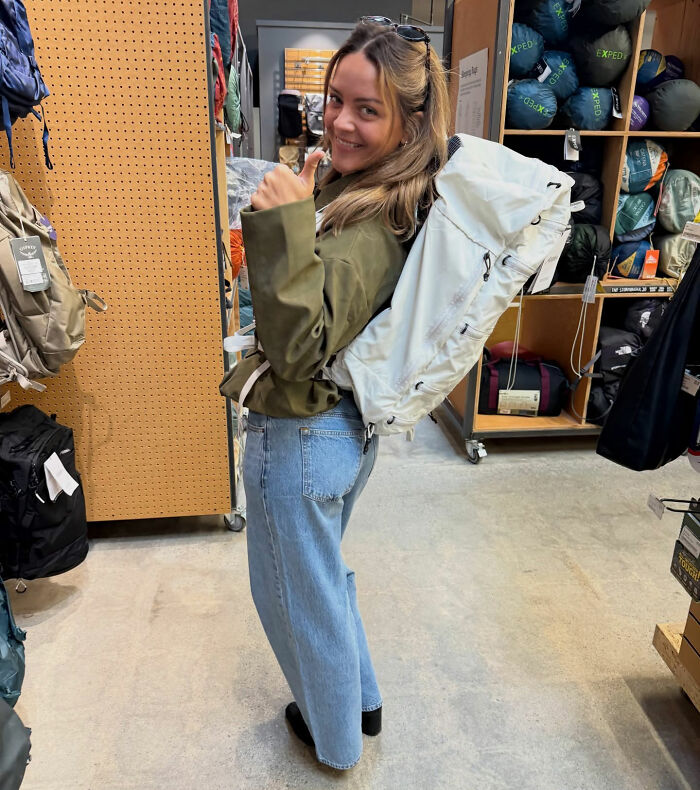 Young woman in casual outfit with large backpack, smiling and giving thumbs up in an outdoor gear store setting.