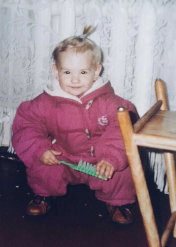 Toddler in a bright pink snowsuit holding a green brush, squatting near a wooden chair in a chaotic childhood photo.