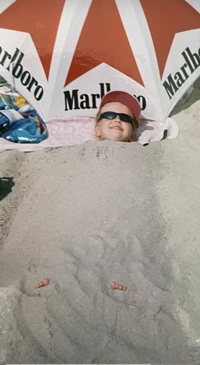 Child buried in sand at the beach under a Marlboro umbrella wearing sunglasses and a pink hat funny chaotic childhood photo.