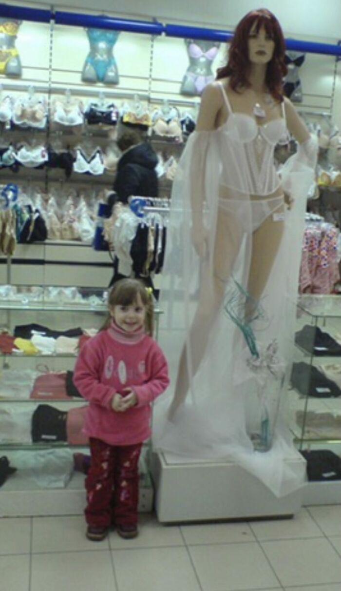 Child smiling next to a mannequin in lingerie inside a store, a funny and chaotic childhood photo moment.