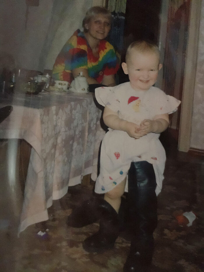 Toddler wearing oversized boots and a dress, smiling in a chaotic and funny childhood photo at home.