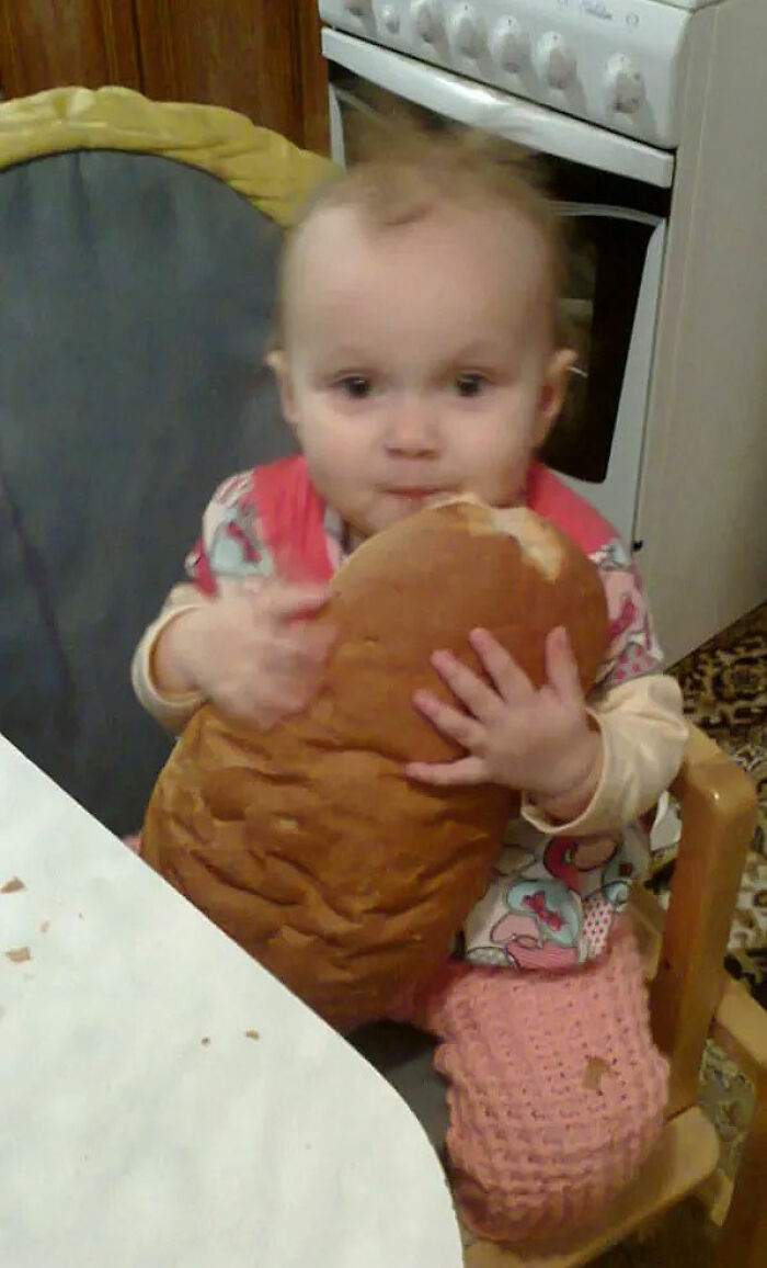 Toddler holding and biting into a large loaf of bread in a chaotic childhood moment at the kitchen table.