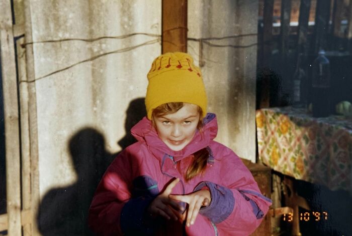 Child in a yellow beanie and pink jacket making a playful hand gesture in a chaotic childhood photo from the 1990s.