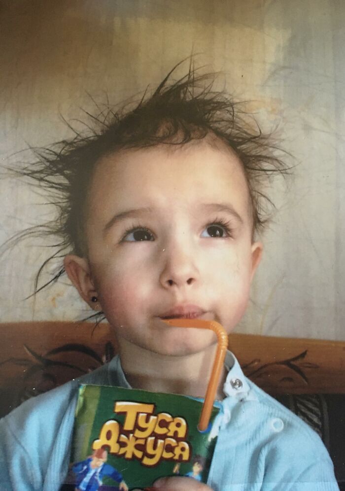 Child with messy hair drinking juice through a straw in a funny and chaotic childhood photo.