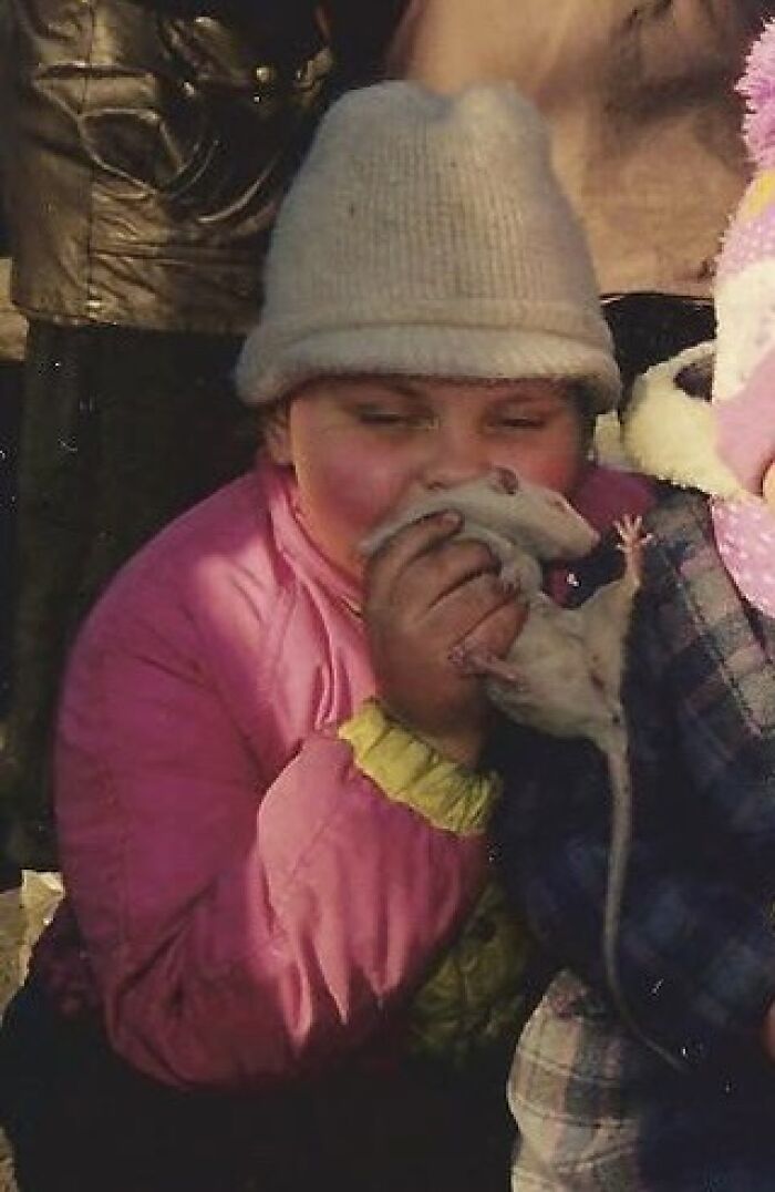 Child holding a rat close to their face, wearing a pink jacket and gray beanie in a funny and chaotic childhood photo.