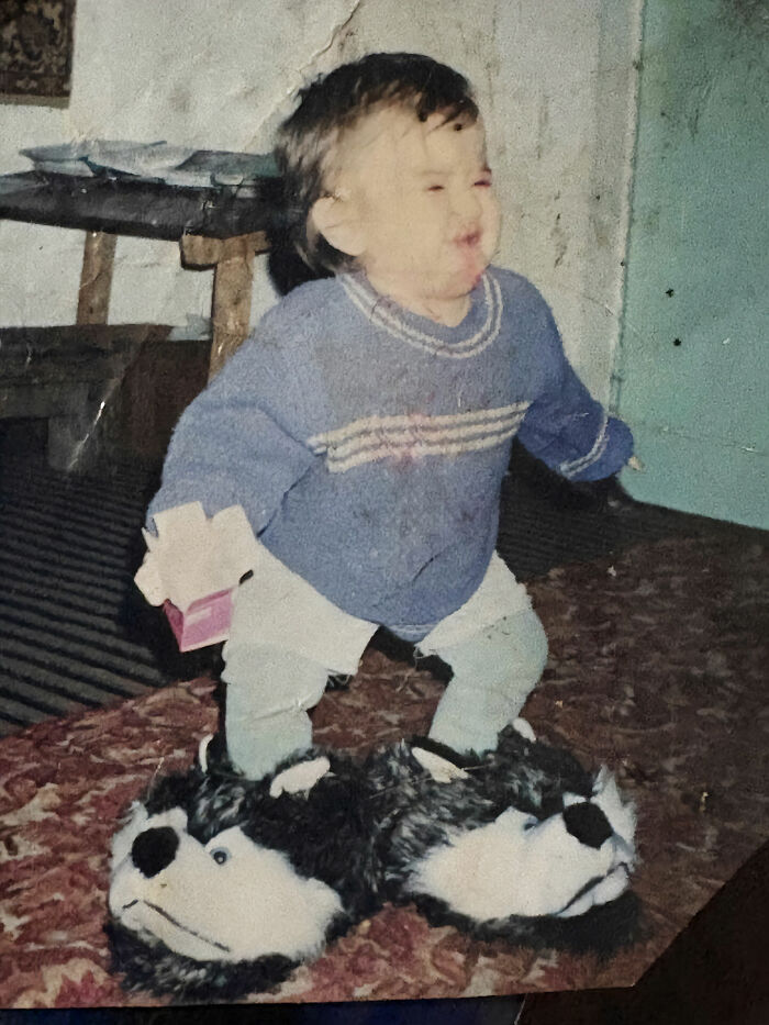 Toddler wearing oversized animal slippers, standing on a patterned carpet in a vintage chaotic childhood photo.