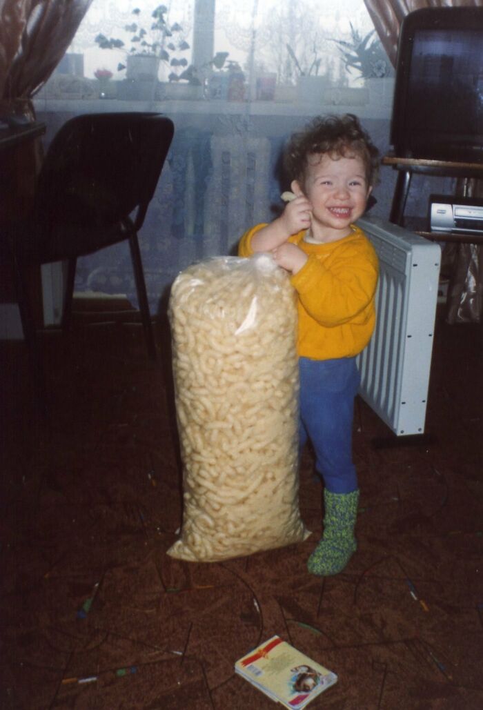 Toddler smiling while hugging an oversized bag of snacks in a chaotic childhood photo at home.