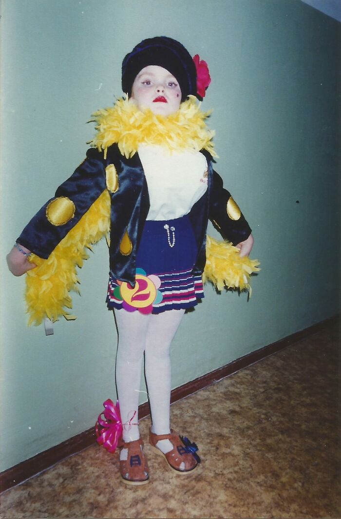 Child in a funny and chaotic childhood photo wearing a colorful, quirky costume with a feather boa and hat confidently posing.