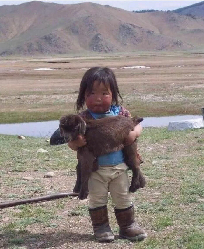 Child holding a goat outdoors in a rural setting, capturing a funny and chaotic childhood moment.