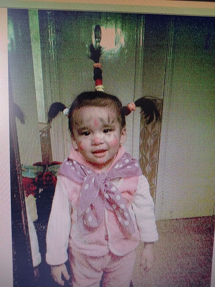Toddler with funny hairstyle and face paint posing indoors in a chaotic childhood photo capturing a humorous moment.