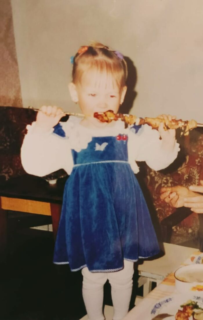 Small child in blue dress eating food on a skewer, a funny and chaotic childhood photo moment captured indoors.