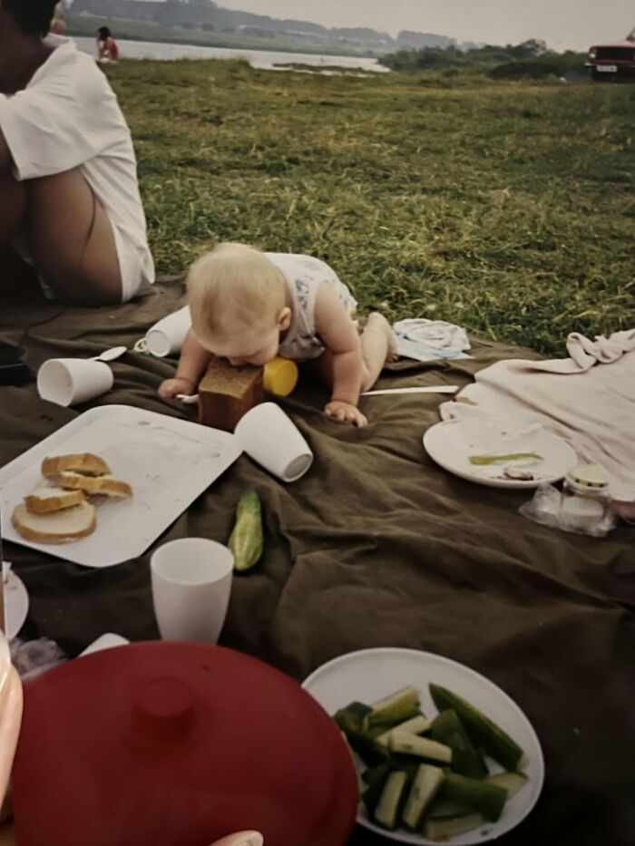 Baby playfully biting a brick during a funny and chaotic childhood picnic outdoors with scattered food and cups.