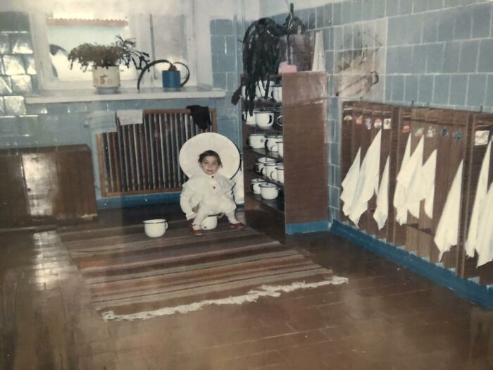 Child in a chaotic childhood photo wearing a white outfit, sitting on a striped rug in a vintage kitchen setting.