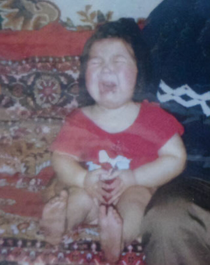 Toddler in a red shirt making a funny chaotic face while sitting on a patterned couch in a childhood photo.