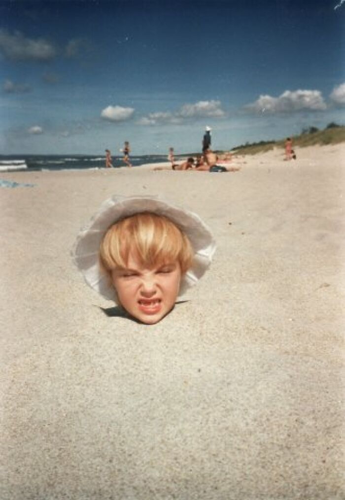 Child buried in sand at the beach with a funny and chaotic childhood expression wearing a white sunhat.