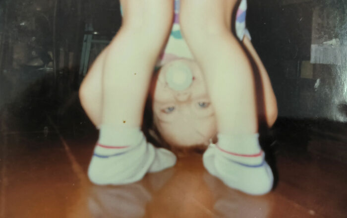 Toddler in socks and a pacifier bending over upside down in a chaotic and funny childhood photo indoors.