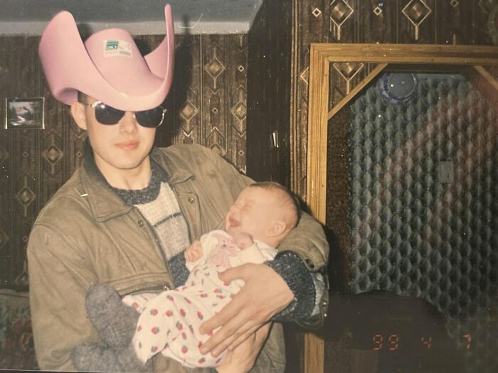Man wearing a pink cowboy hat and sunglasses holding a crying baby in a chaotic childhood photo from the 90s.