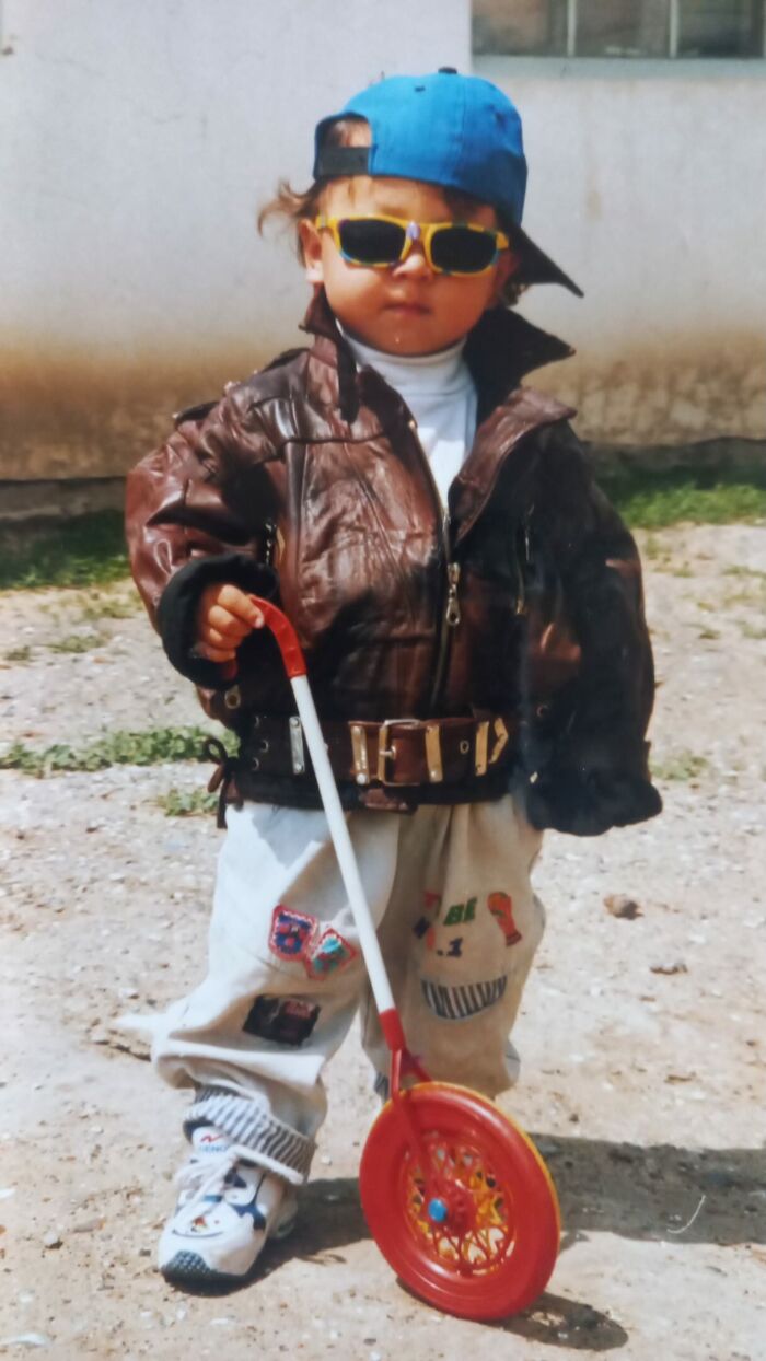 Toddler in sunglasses, leather jacket, and cap holding a red toy wheel in a funny and chaotic childhood photo outdoors.