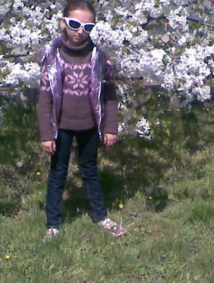 Child wearing large sunglasses and patterned sweater standing awkwardly in front of blooming tree in funny chaotic childhood photo