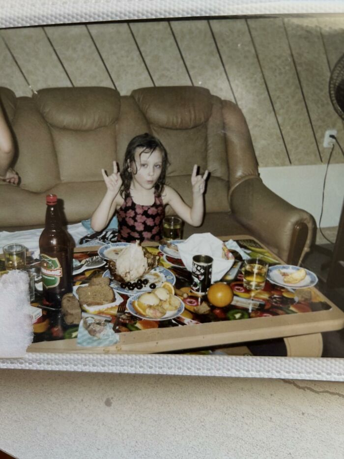 Child making funny hand signs at a cluttered table with food and drinks in a chaotic childhood photo setting.